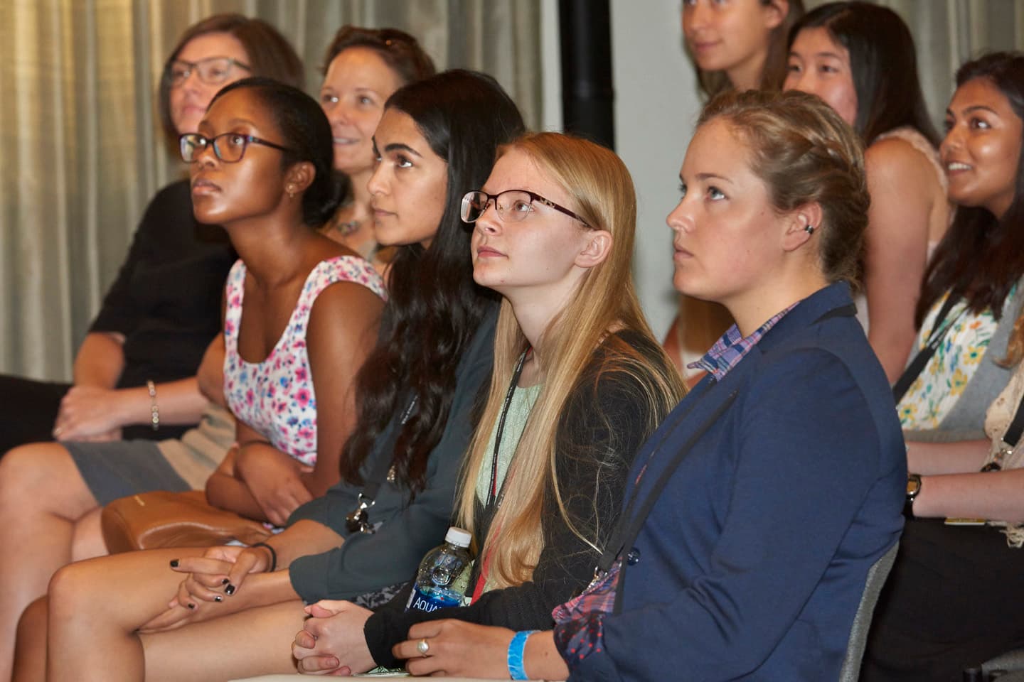 A row of four girls sit in a row and listen to a lecture. The speaker is not pictured.