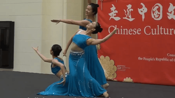 Three dancers in traditional Chinese folk attire perform on a stage in front of a red banner reading “Chinese Culture Day.”