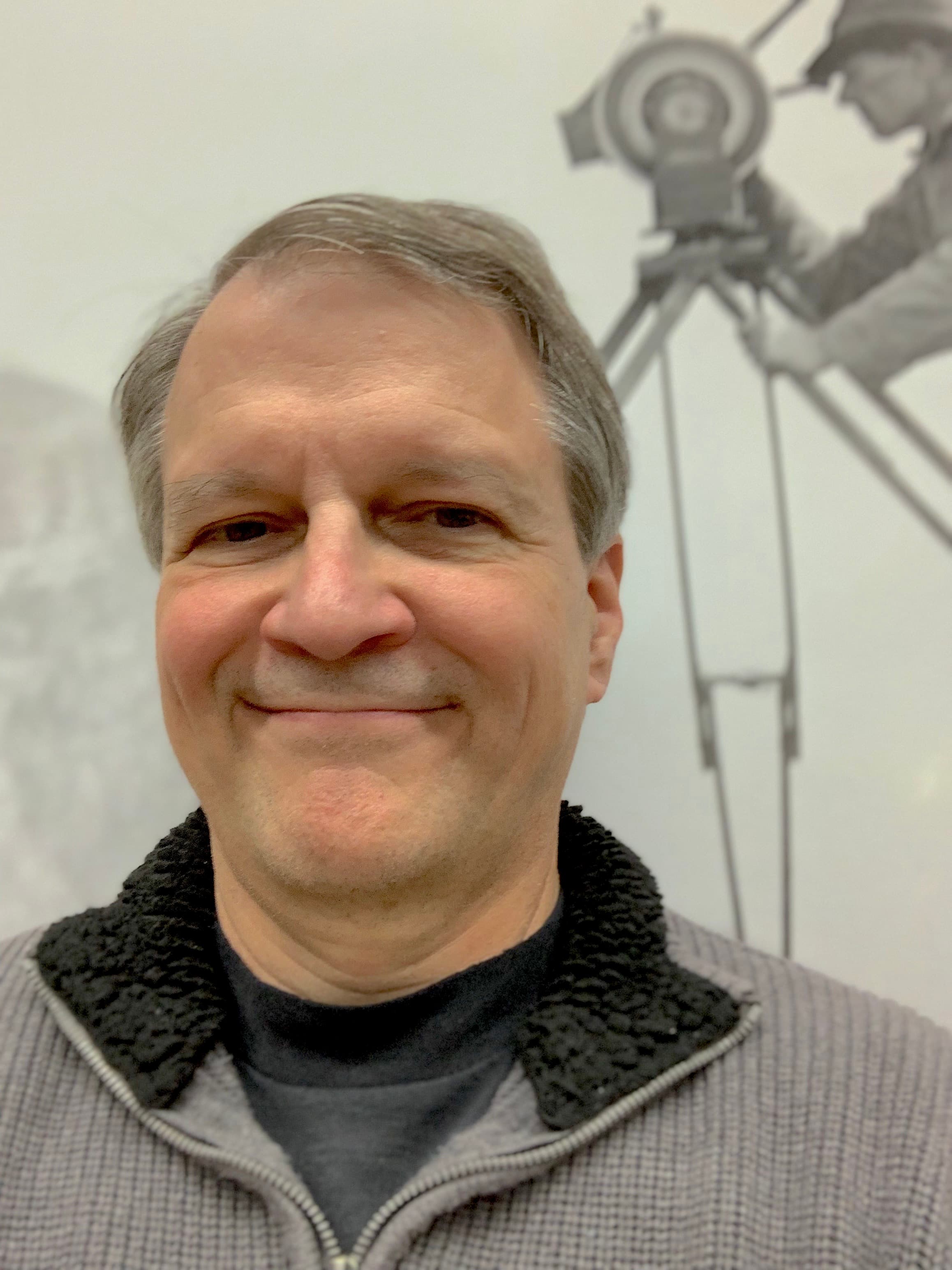 A smiling man with short gray hair, standing in front of an archival picture of a man using an old cinema-camera on a tripod.