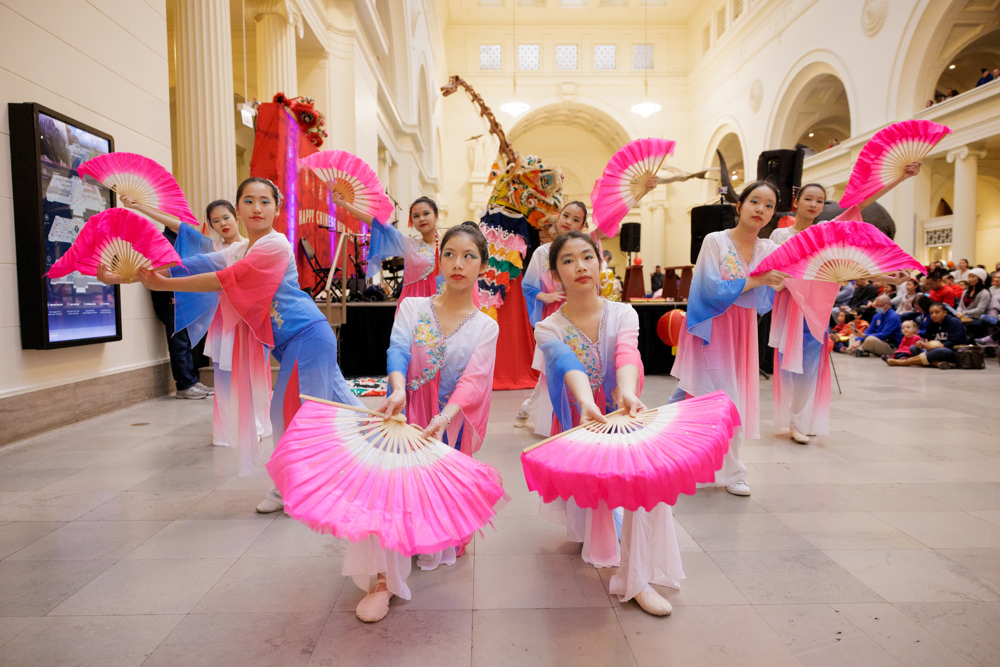 Dancers in traditional dress pose holding pink hand fans in the large main hall of the museum.