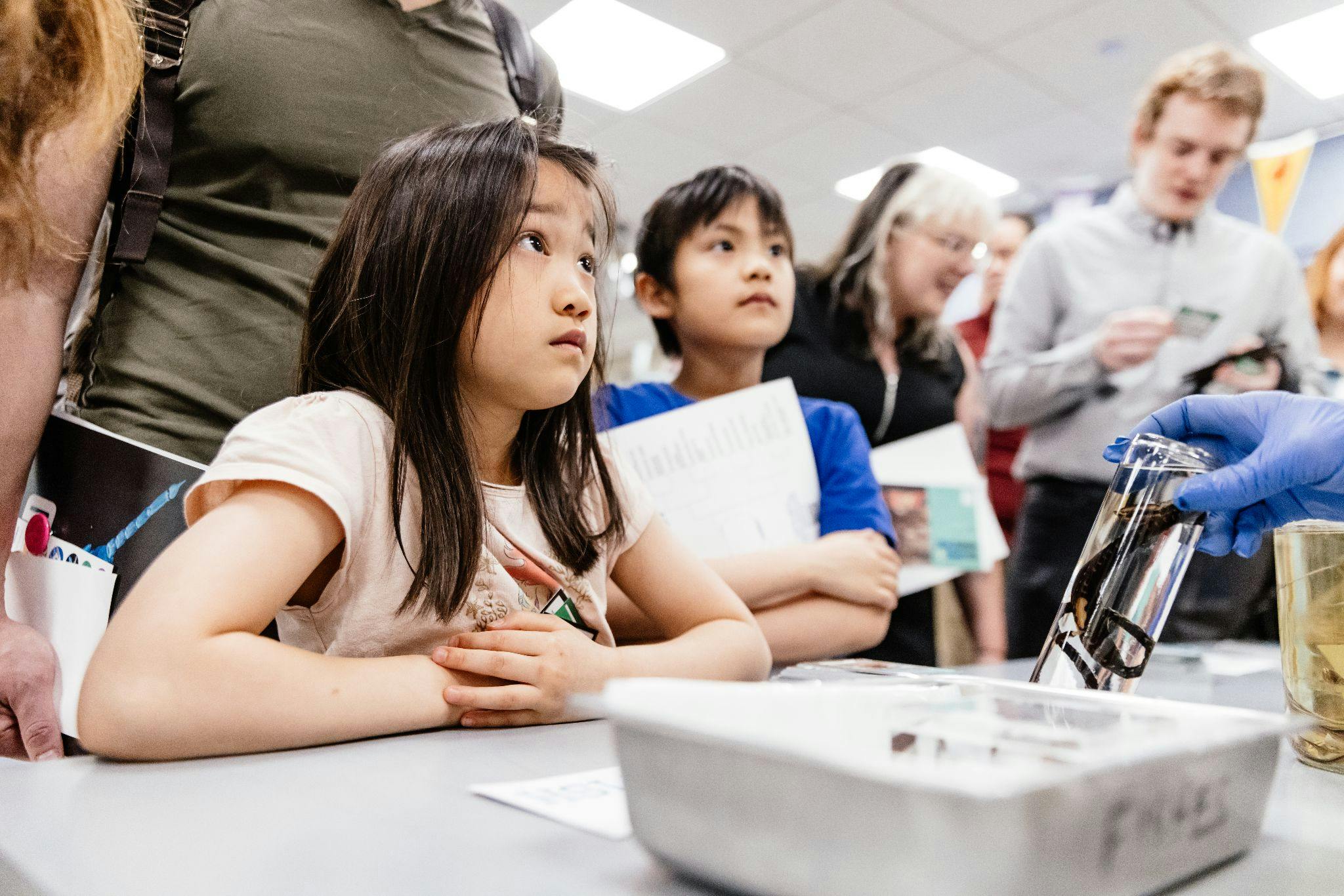 Two young children look up at a person giving a demonstration. Several other people can be seen in the background gathered around.