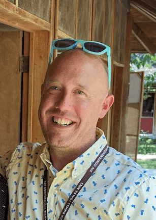 A man, smiling and wearing a button down shirt. His sunglasses are perched on top of his head.