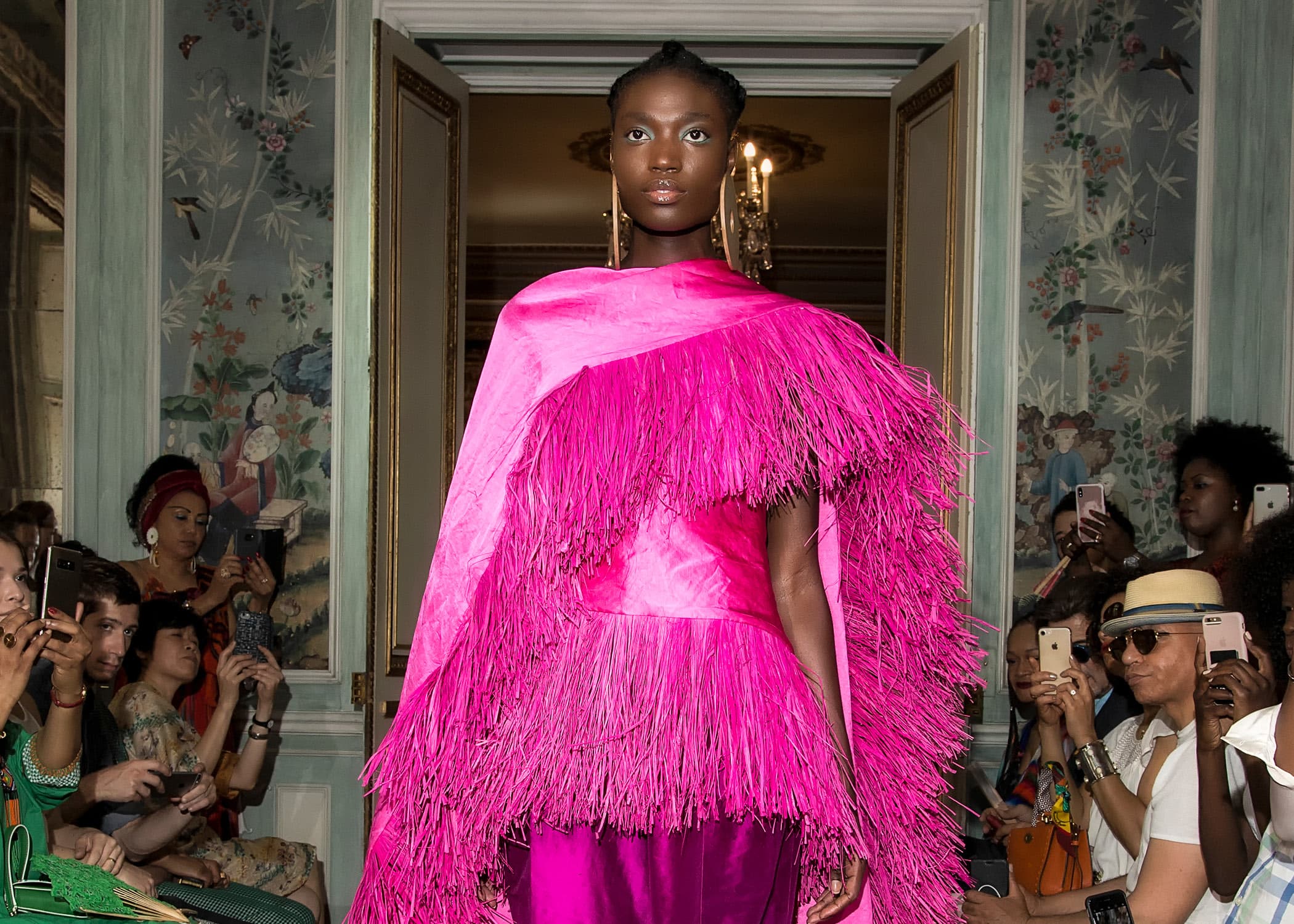 A model wearing a bright pink, fringed ensemble walks through a dooorway. People on either side of her are holding phones up as if recording or taking pictures.