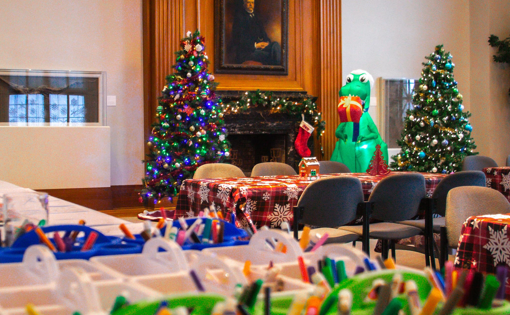 A room with a fireplace, decorated for Christmas, with two lighted trees, garland on the mantle, and an inflatable dinosaur.