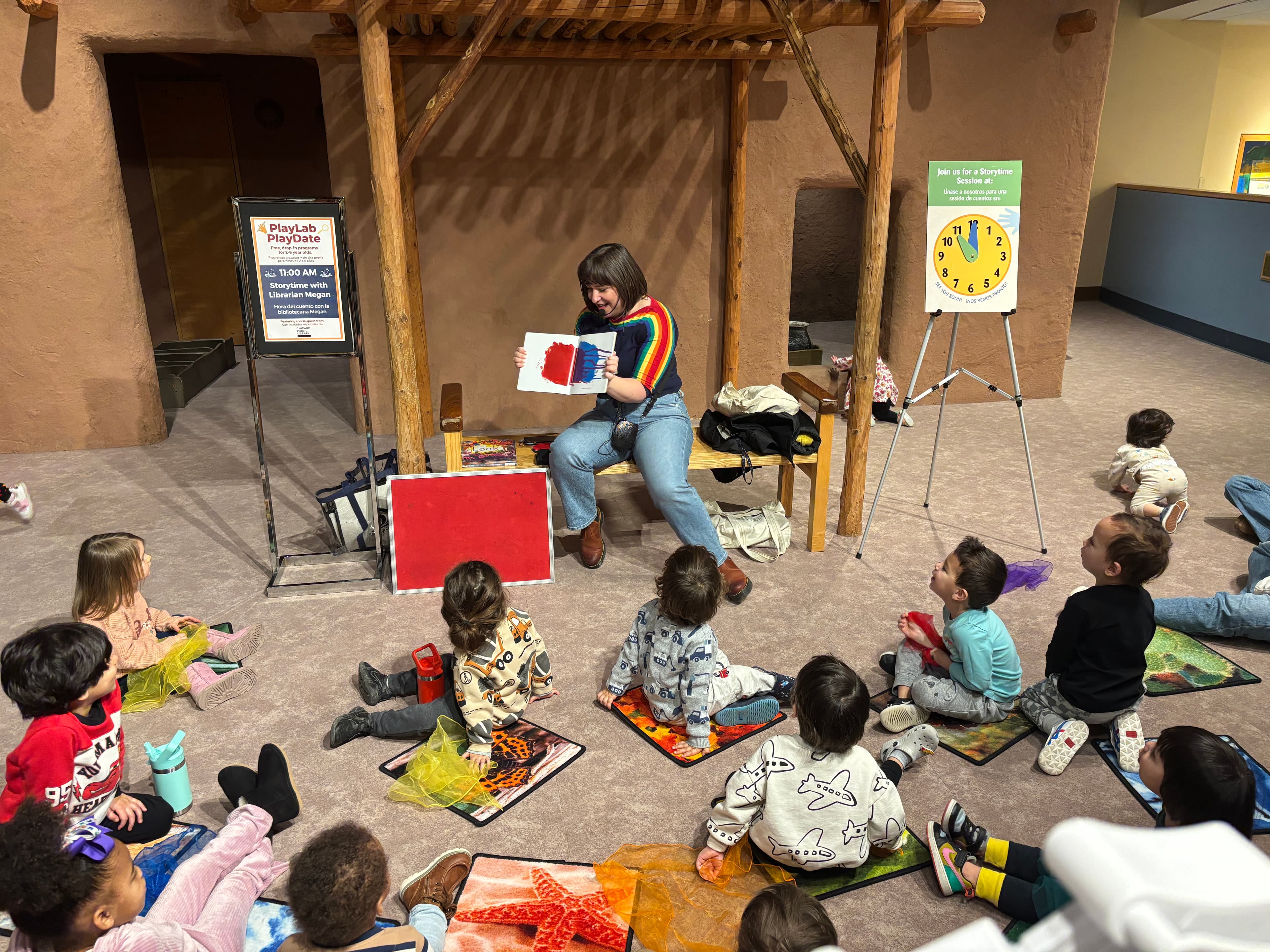 A women sits on a bench holding a book, facing a group of children seated on mats.